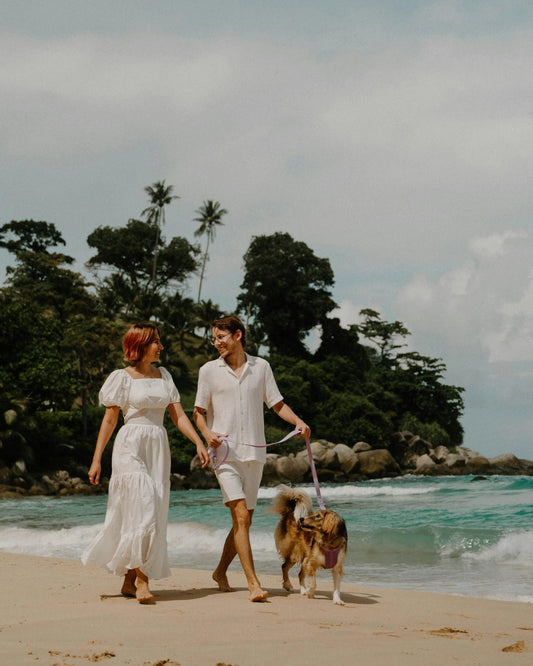 Couple souriant promenant un chien en laisse rose sur une plage, sous un ciel ensoleillé